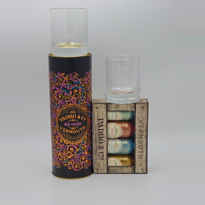 A gift set featuring a tall black cylindrical tin with ornate multicolor scrollwork and a clear glass on top, next to a rustic wooden crate labeled 'PADRO & CO VERMOUTH' containing several small bottles with colorful labels.