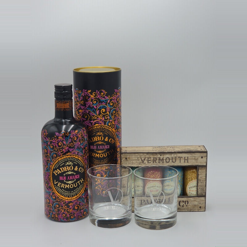 A bottle of Padró & Co Vermouth with a matching cylindrical box, two clear engraved whiskey glasses, and a small wooden crate labeled Vermouth, arranged on a light gray studio background.