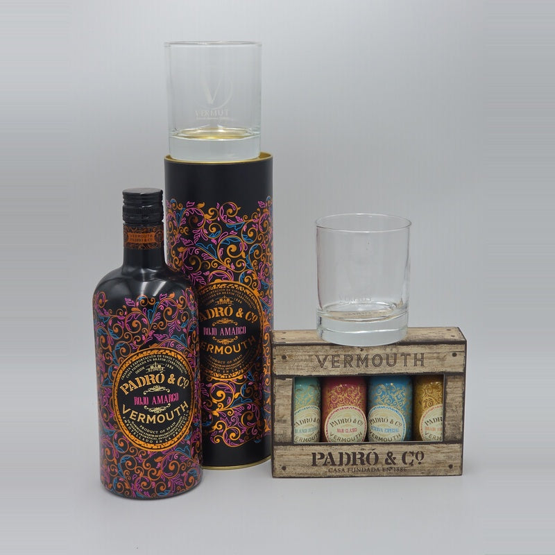 A bottle of Padró & Co Vermouth with a matching ornate black cylinder box, a clear glass, and a small wooden crate labeled VERMOUTH containing four colorful bottles, photographed on a light gray studio background.