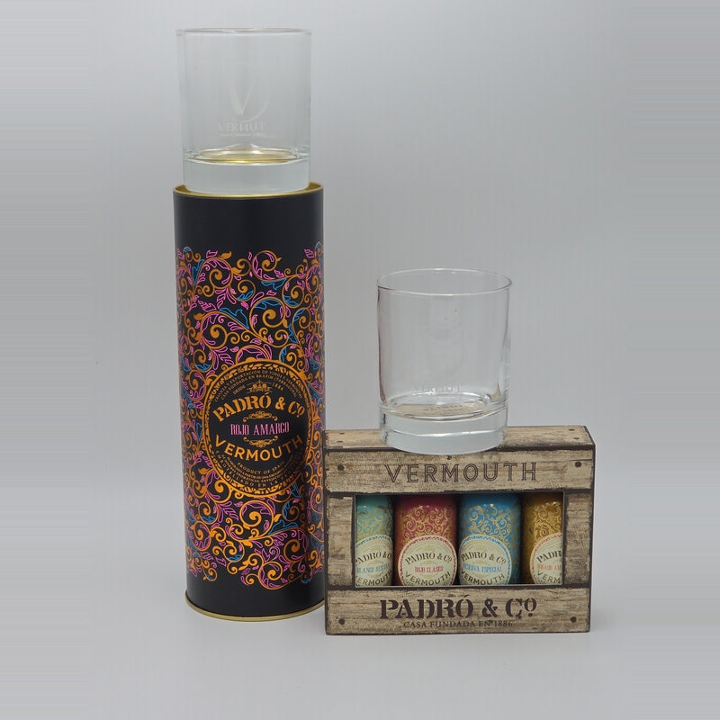 A Pedro & Co Vermouth gift set featuring a tall black cylindrical tin with ornate multicolor scrollwork and a clear glass, alongside a small wooden crate labeled 'VERMOUTH PADRO & CO' containing several colorful bottles and a clear glass.