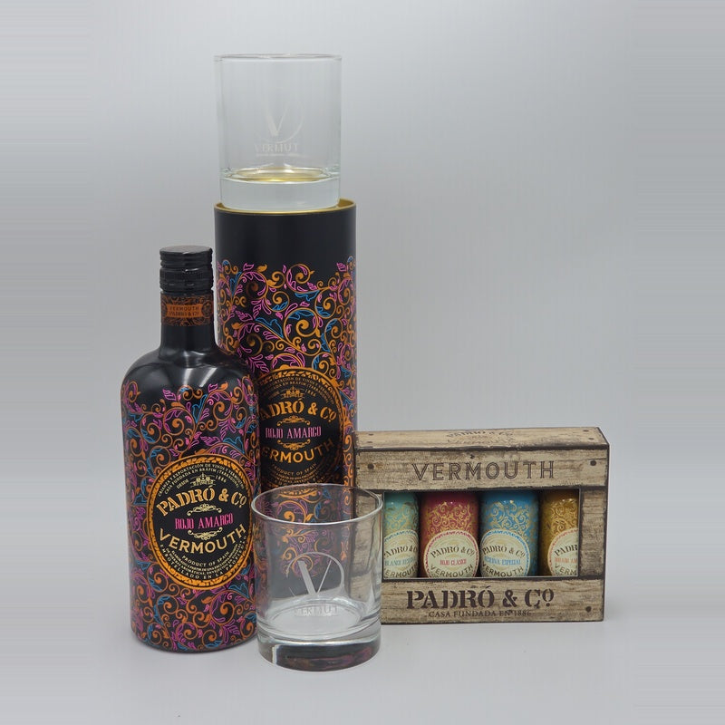 A bottle of Padró & Co Vermouth with a matching patterned cylinder, a clear glass, and a wooden crate box containing five smaller bottles, photographed on a light gray studio background.