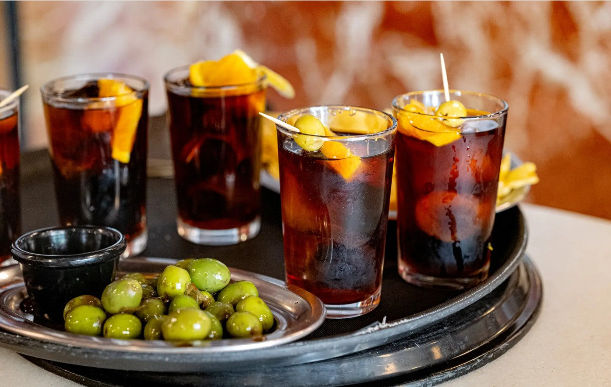 Four glasses of dark liquid with garnishes on a tray, accompanied by olives.